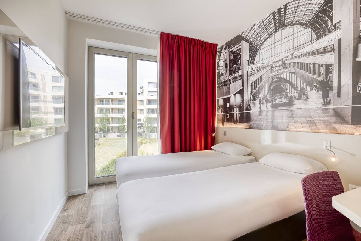 A hotel room with a bed, a desk, a chair, a window with a red curtain, and a wall-mounted TV.
