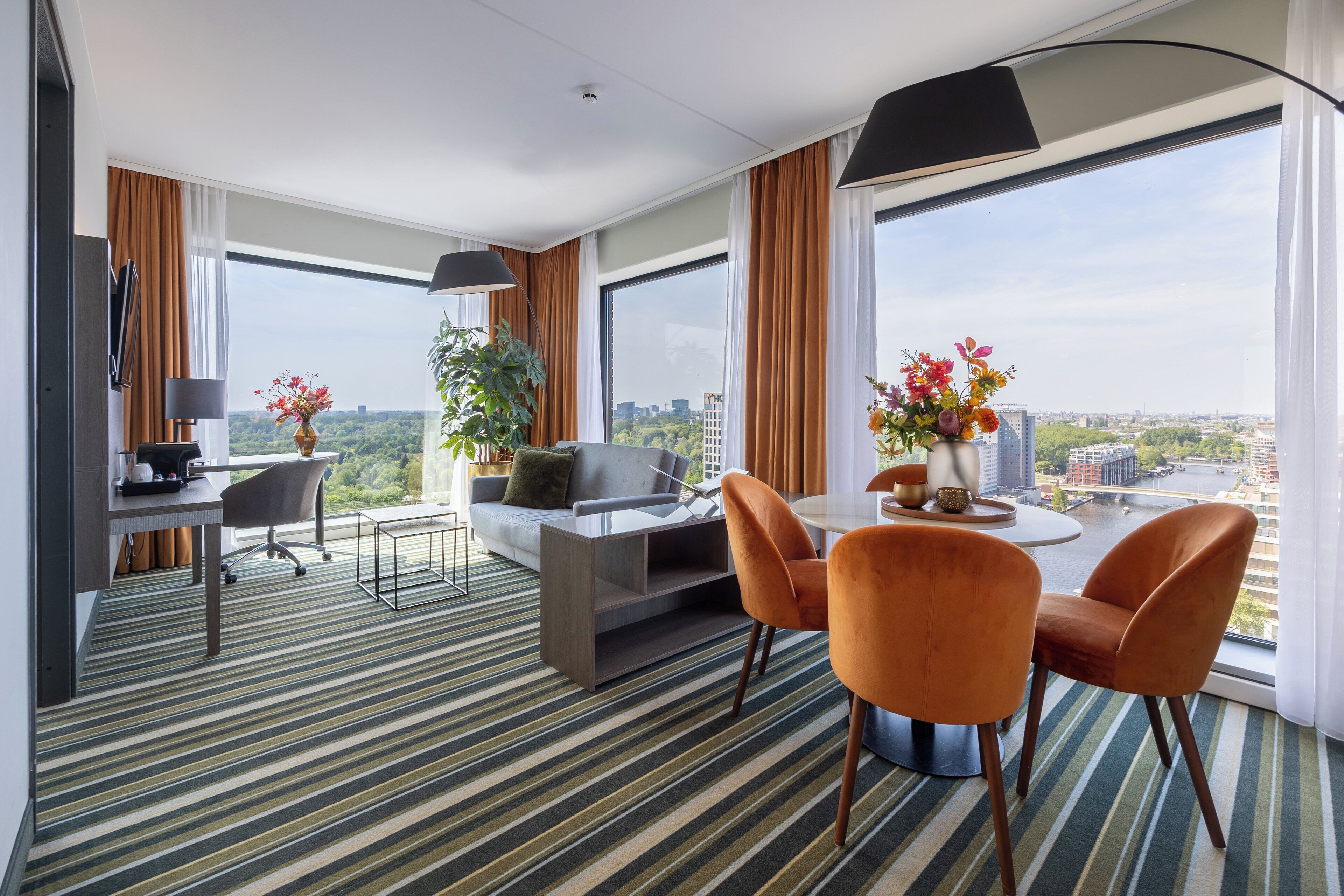A modern hotel room with a dining area, a sofa, a TV, and a large window with a city view.
