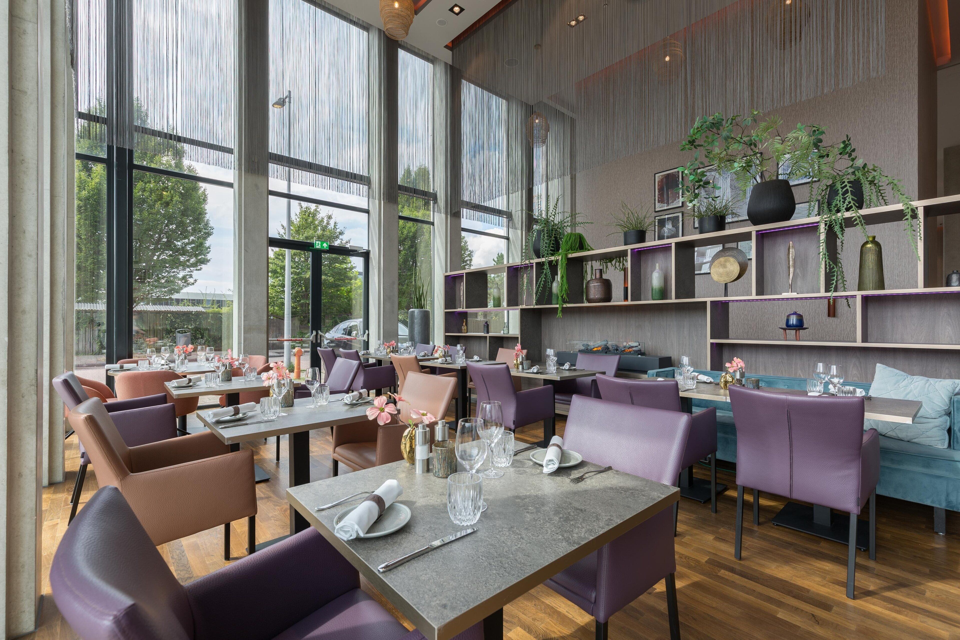 A modern restaurant with large windows, purple seating, and a variety of plants on shelves.