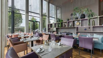 En modern restaurang med stora fönster, lila sittmöbler och ett stort utbud av växter i hyllor.