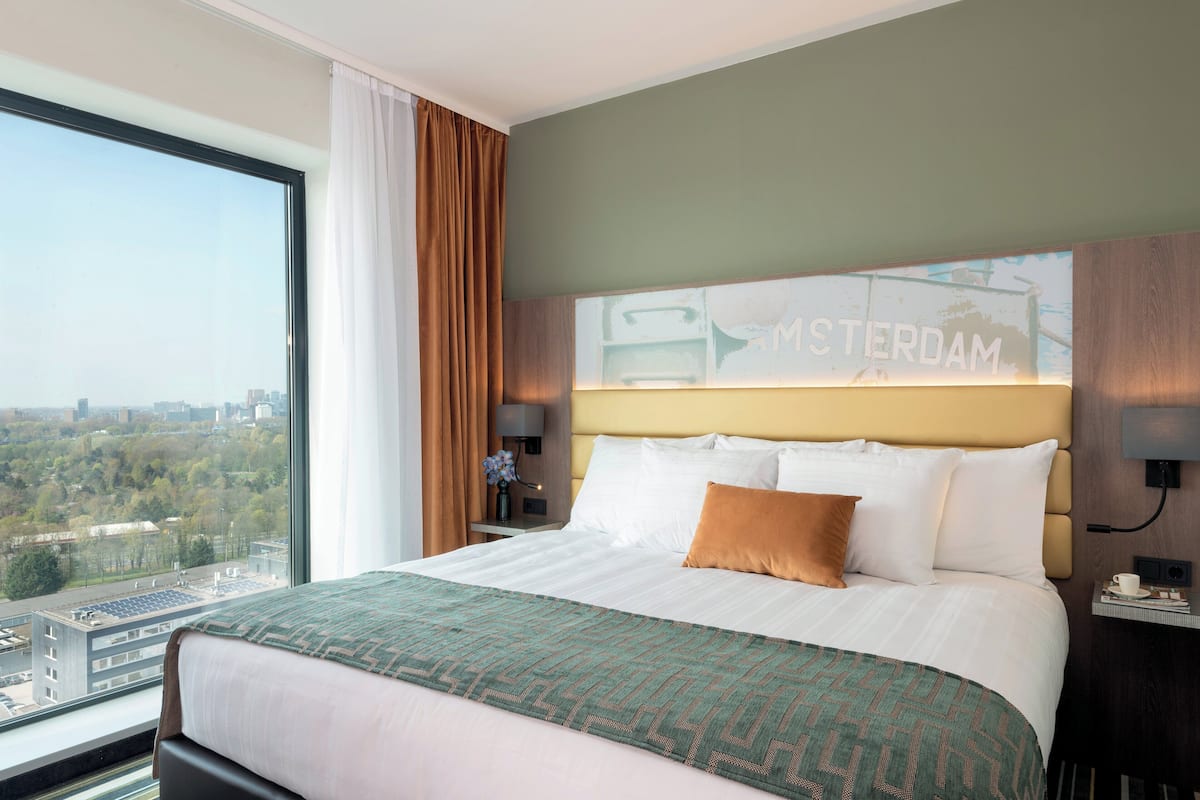 A hotel room with a large bed, a view of the city, and a sign on the wall that says "MESTERDAM".