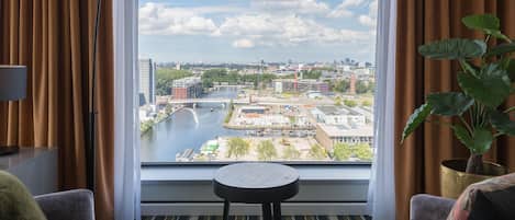 A hotel room with a large window offering a view of a cityscape, including a river and buildings. There are two chairs and a small table in the room.