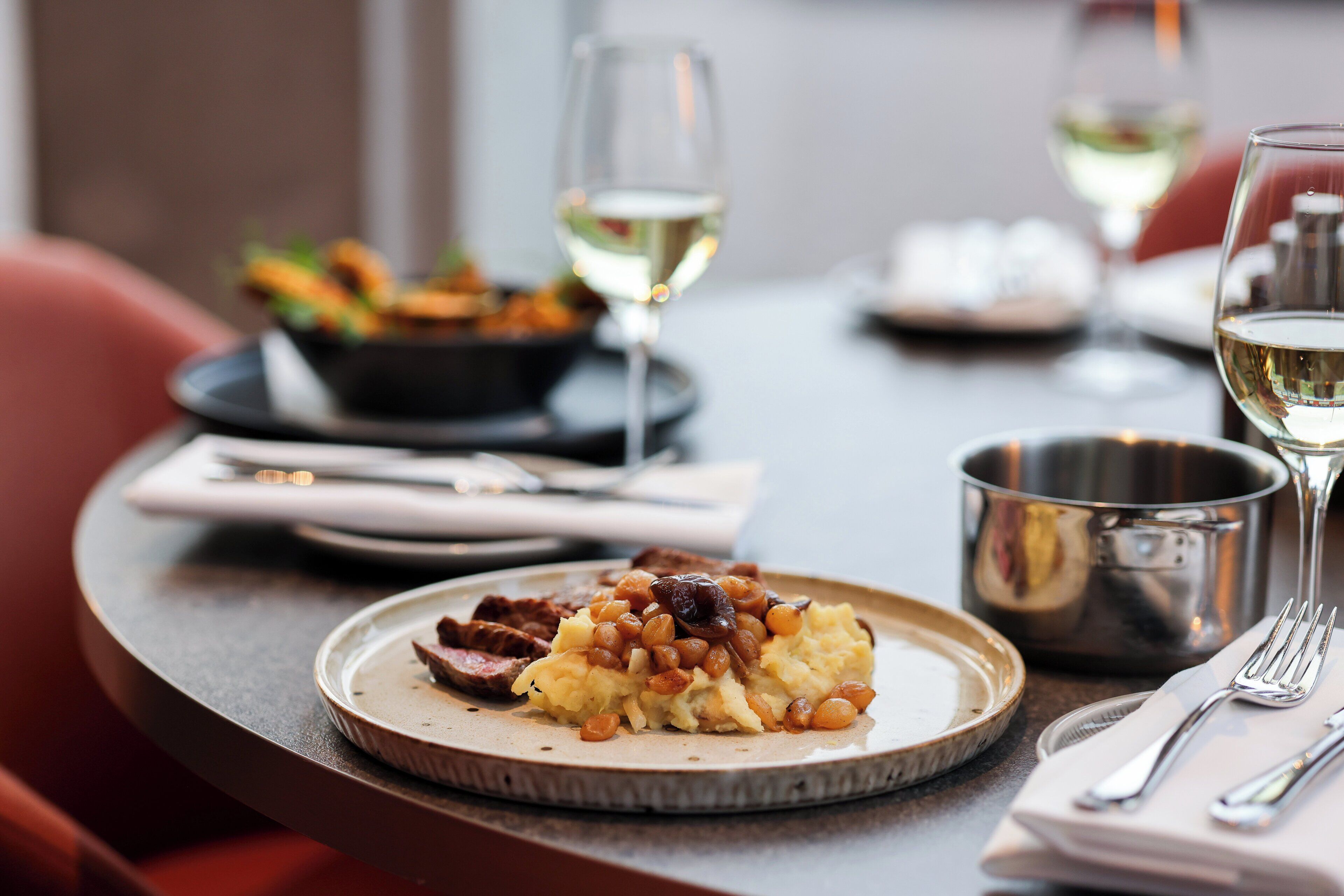 A dining table with a plate of food, a glass of white wine, and a metal pot.
