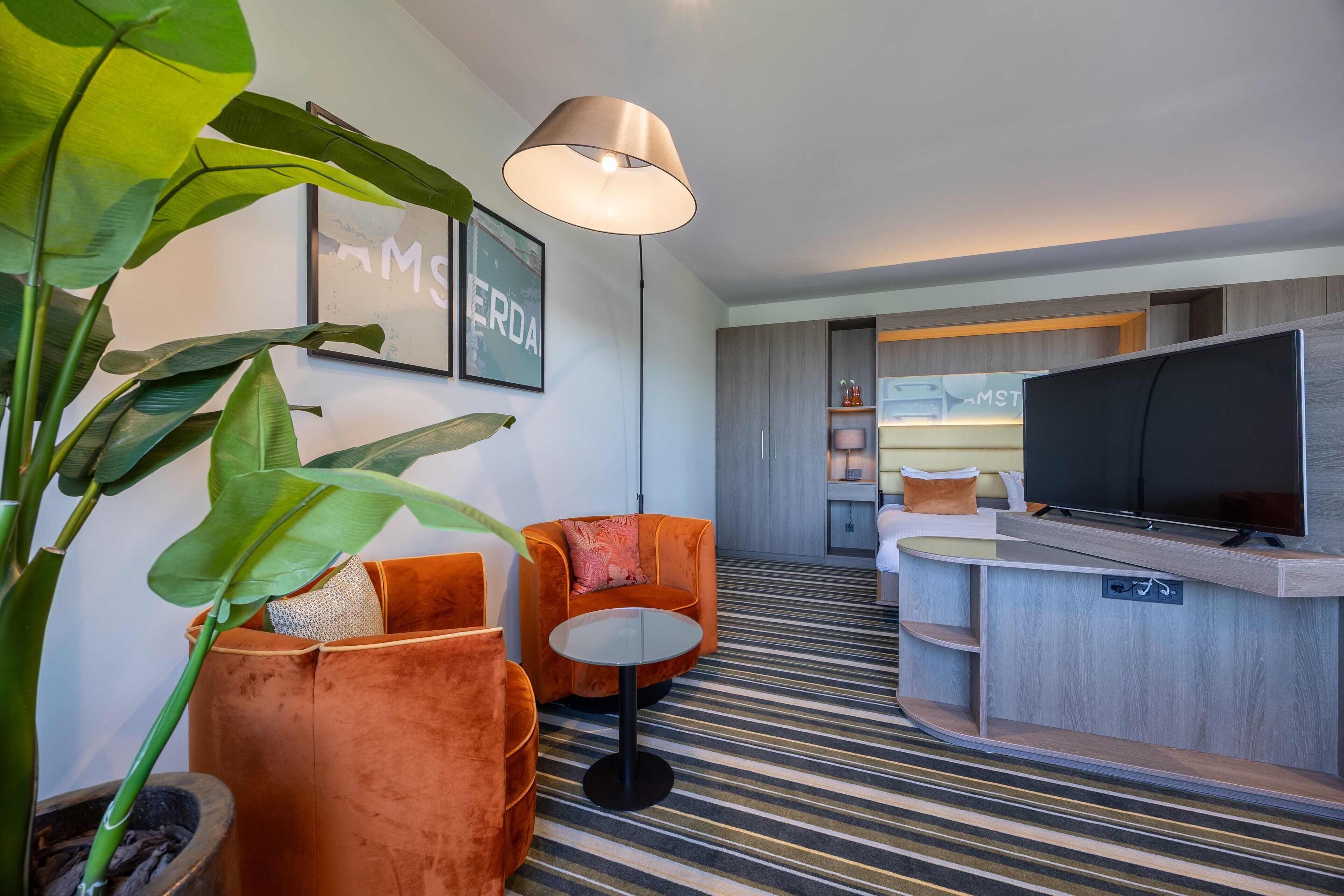 A modern hotel room with a striped carpet, orange armchairs, a round table, a large TV, and a pendant light.