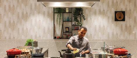 Un cocinero preparando comida en una cocina moderna con una estufa grande, diversos utensilios de cocina y una pared decorativa.