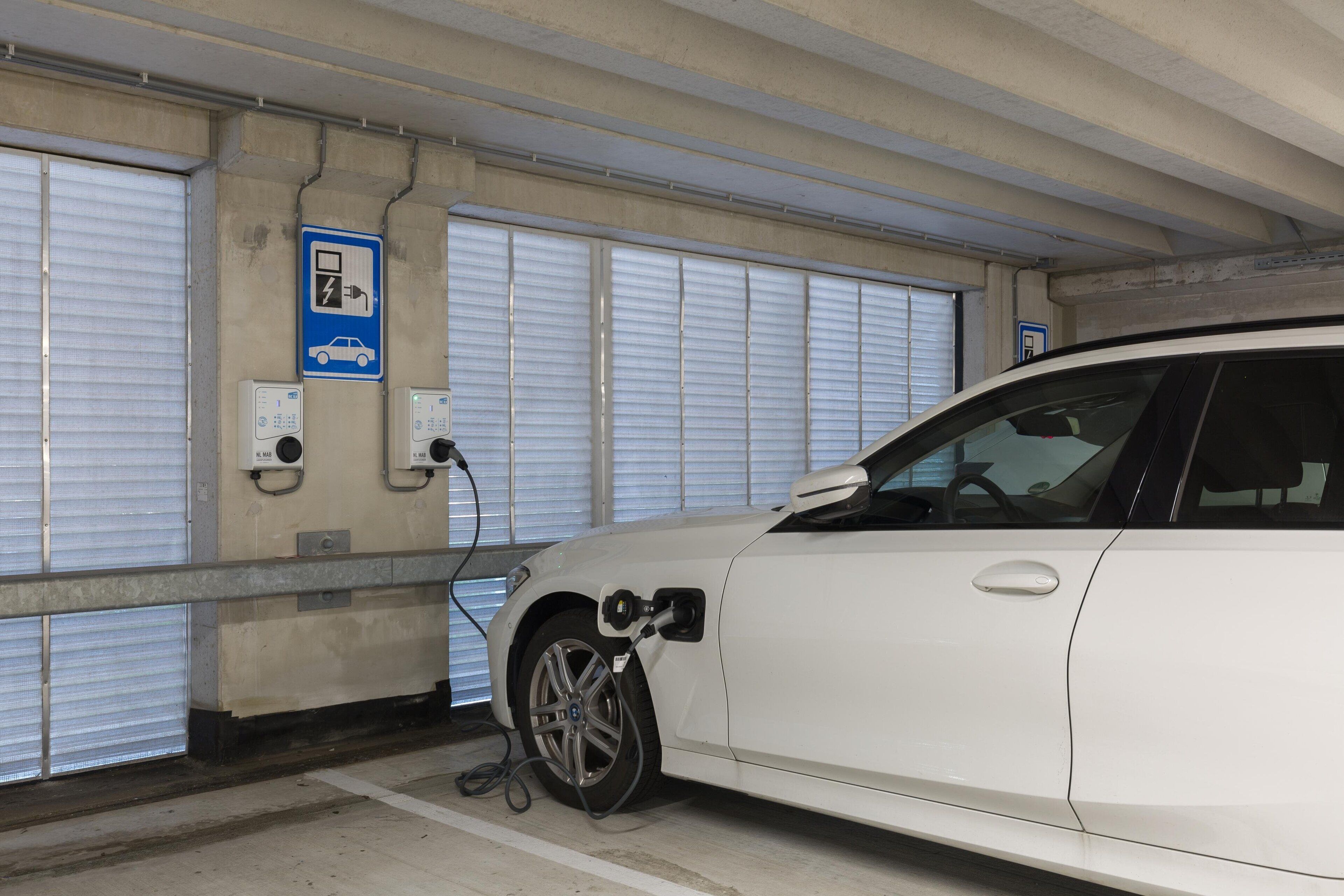 Un coche blanco aparcado en una zona de aparcamiento cubierta, con estación de carga y señal azul que indica carga para vehículos eléctricos.