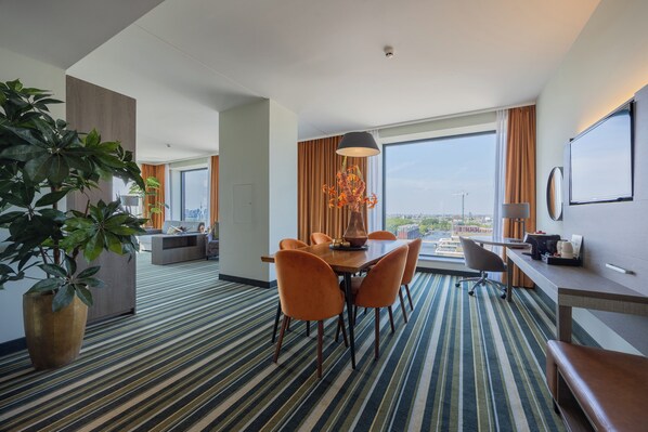 A hotel room with a dining area, a large window with a view, a potted plant, and a modern lamp.