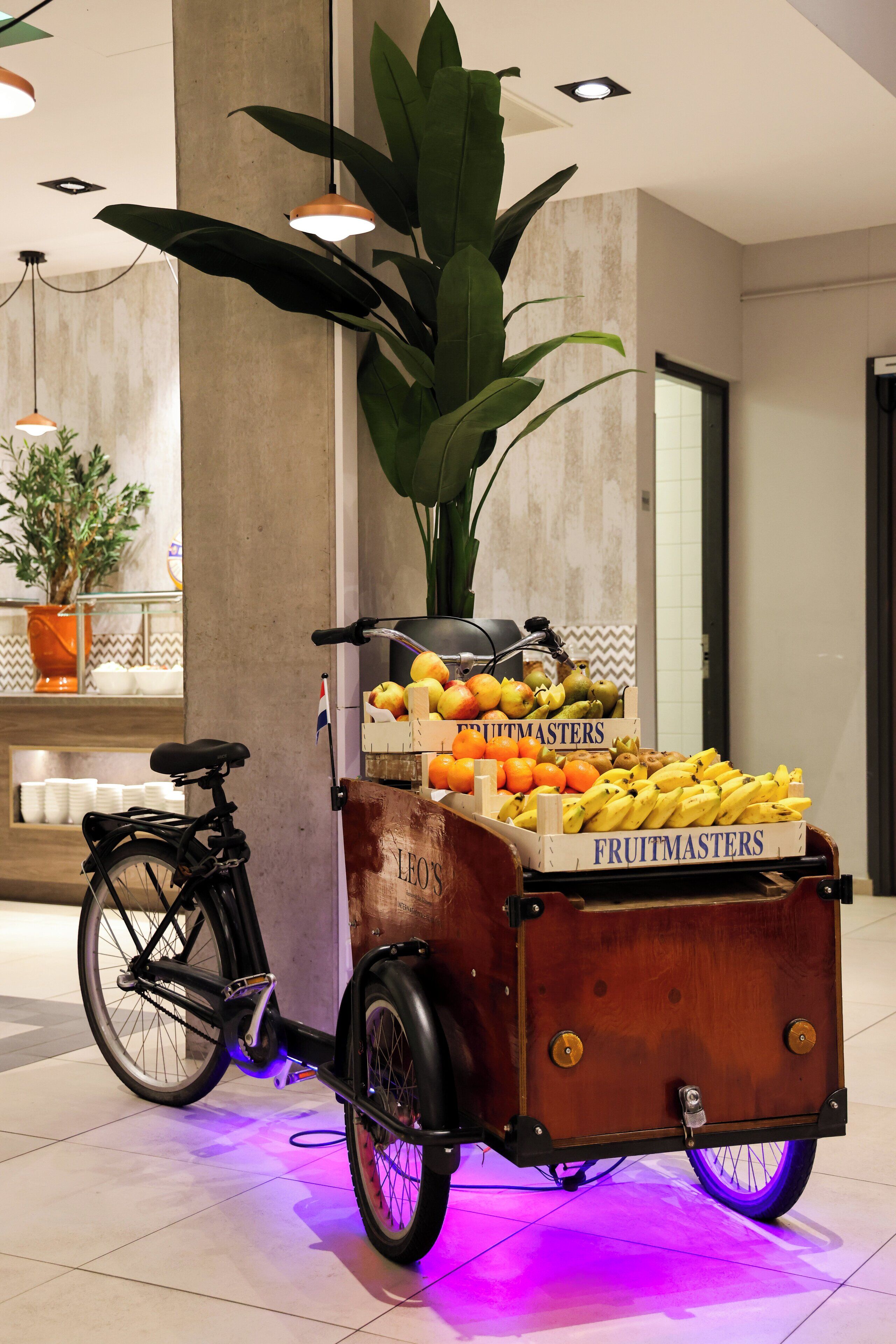 A stationary bicycle cart with a fruit display, a potted plant, and a bicycle parked in a lobby area.