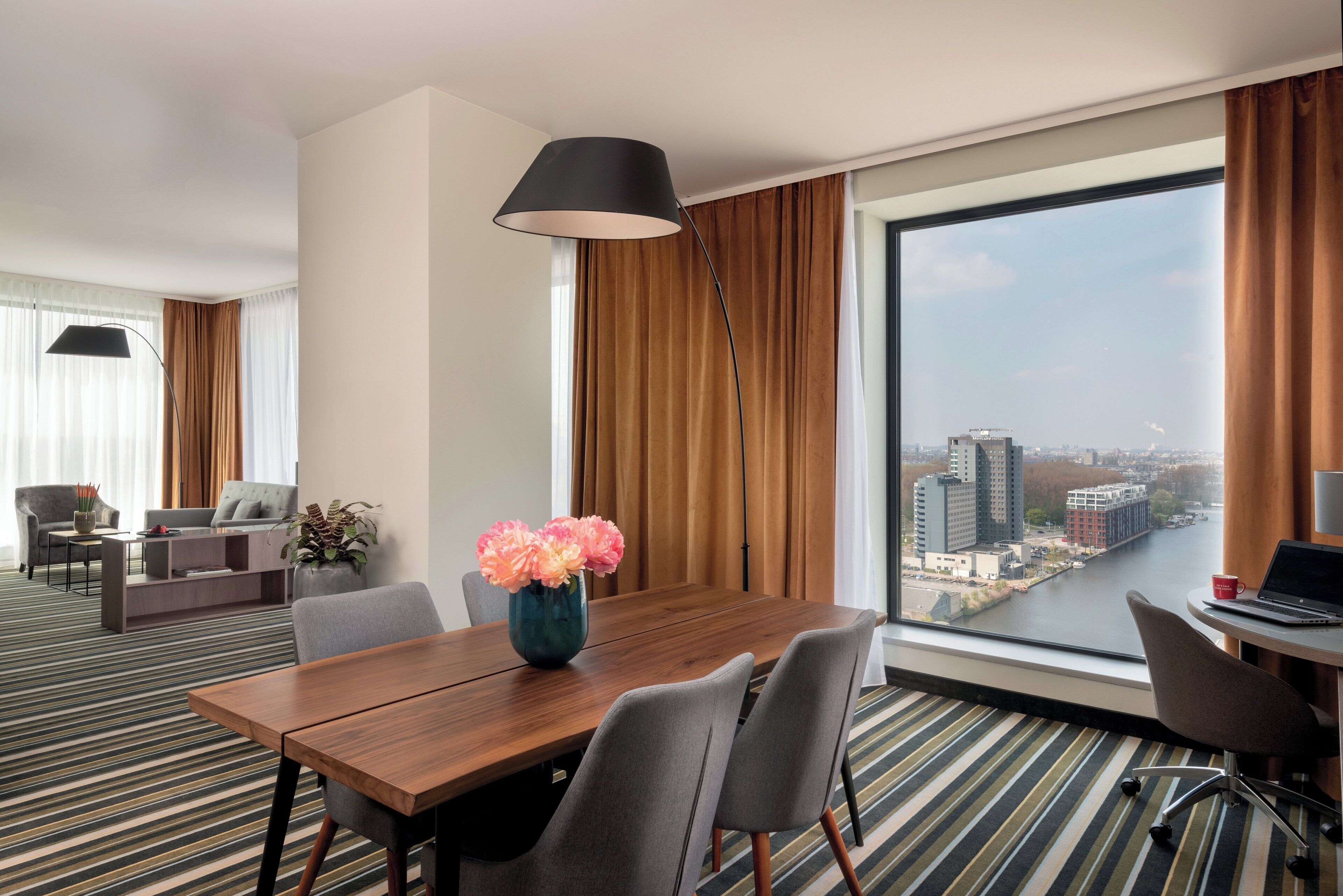 Een moderne hotelkamer met een groot raam, een houten eettafel, grijze stoelen, een zwarte vloerlamp en uitzicht op een rivier en stadsbeeld.
