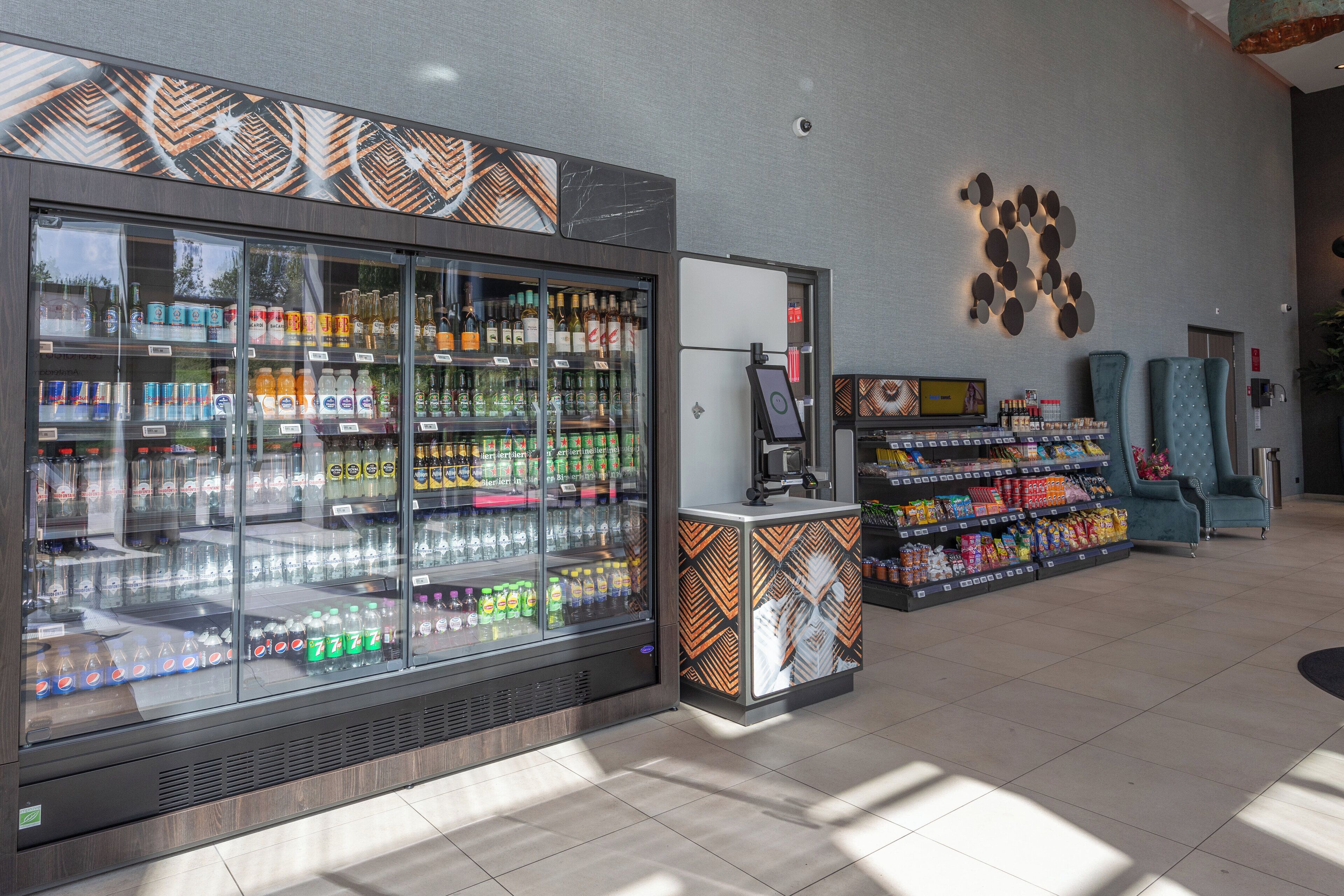 Un área comercial moderna con mostradores refrigerados para bebidas, una estación de autopago y zonas de estar.