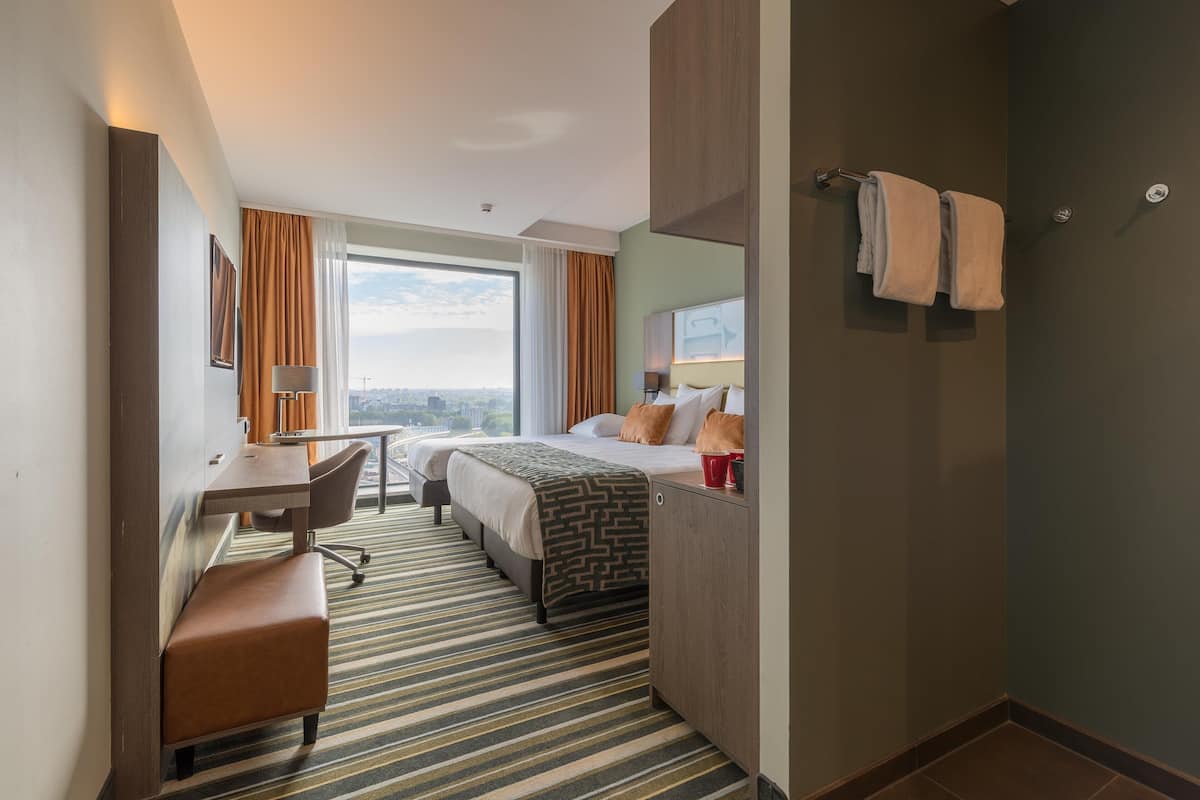 A hotel room with a bed, desk, chair, and a view of the city.