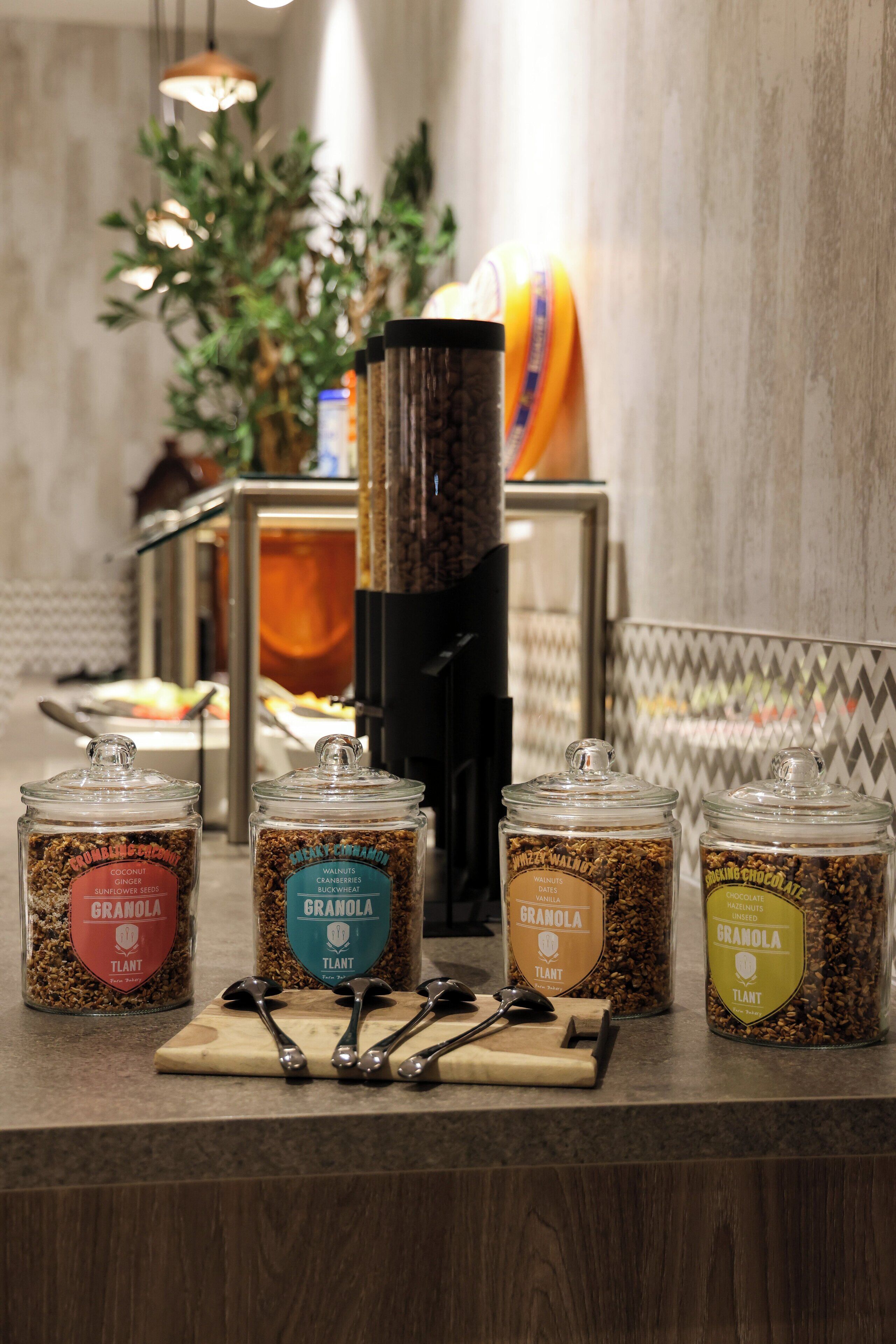 Ein Frühstücksbuffet mit Gläsern voller Granola in verschiedenen Geschmacksrichtungen wie Kokos und Schokolade, eine Kaffeemaschine und eine Obstschale.