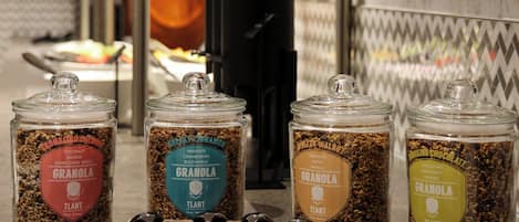 A breakfast bar with granola jars labeled with flavors like coconut and chocolate, a coffee maker, and a fruit bowl.