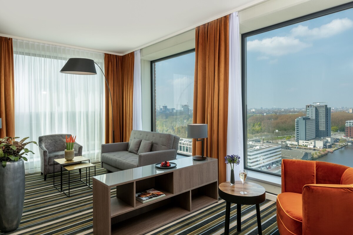 A modern hotel room with a large window offering a city view, a sofa, armchairs, a coffee table, and a lamp.