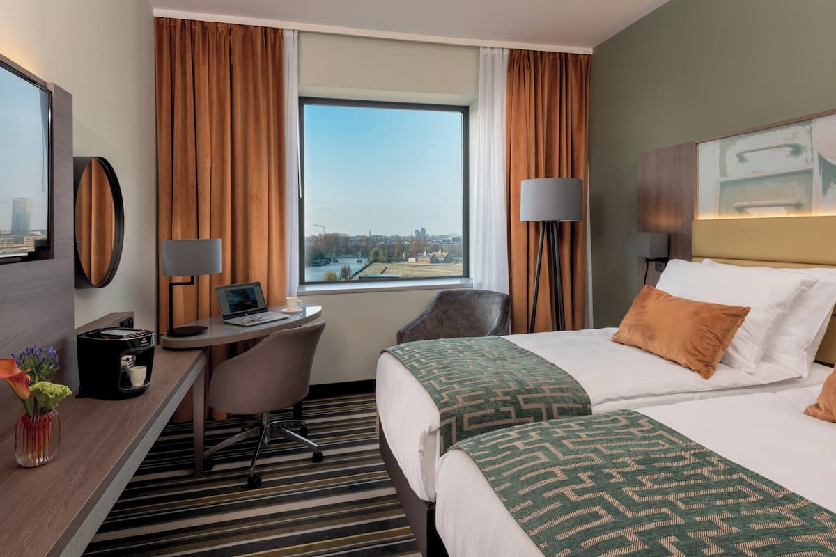 A hotel room with a large bed, a desk with a laptop, a chair, and a view of a cityscape through a window.
