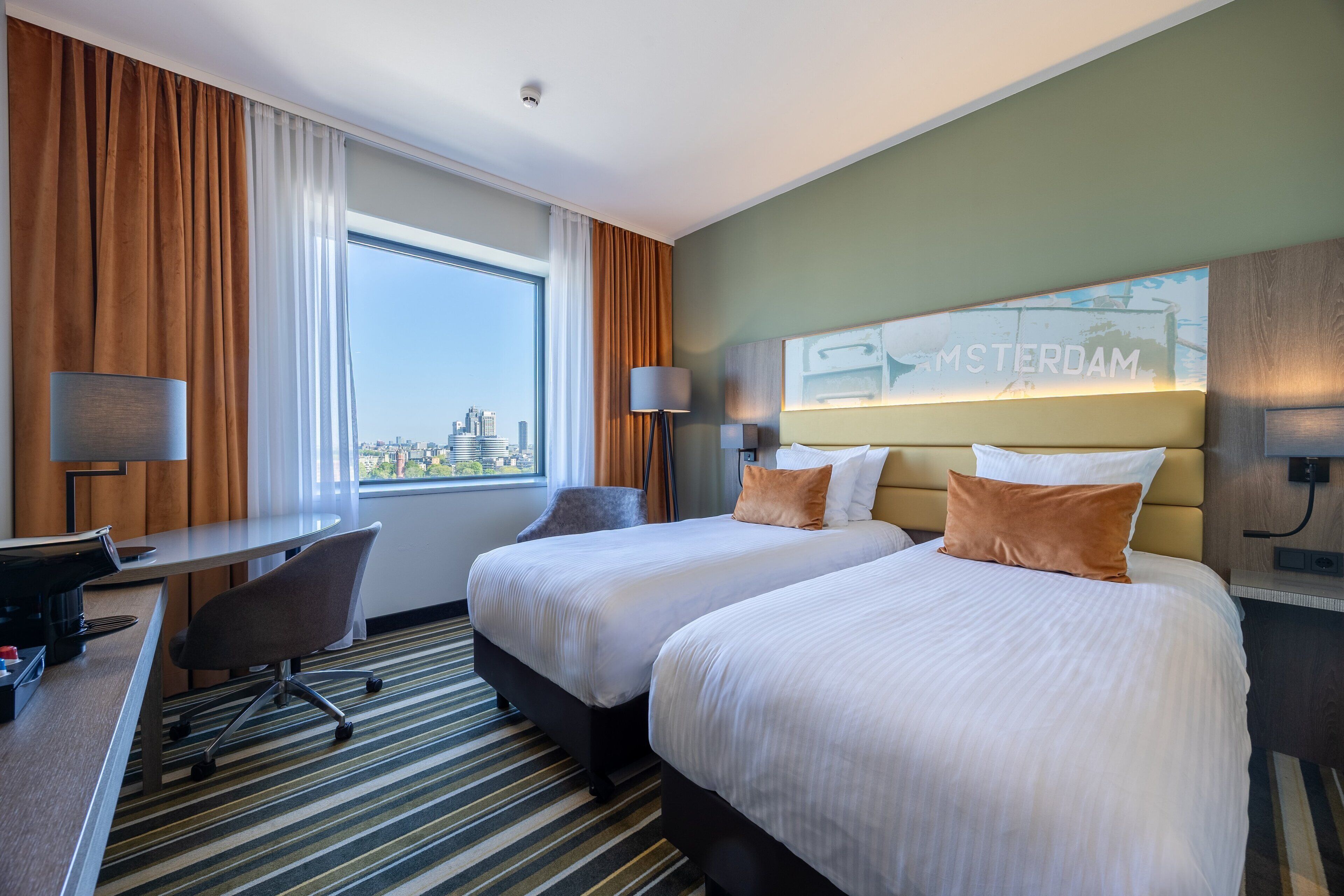 A hotel room with two beds, a desk, and a chair. There is a view of the city through the window.