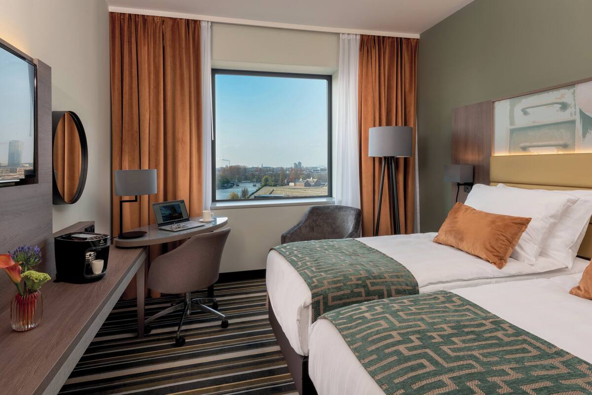 Camera d'albergo con un letto grande, una scrivania con un portatile, una sedia e vista sulla città attraverso una finestra.