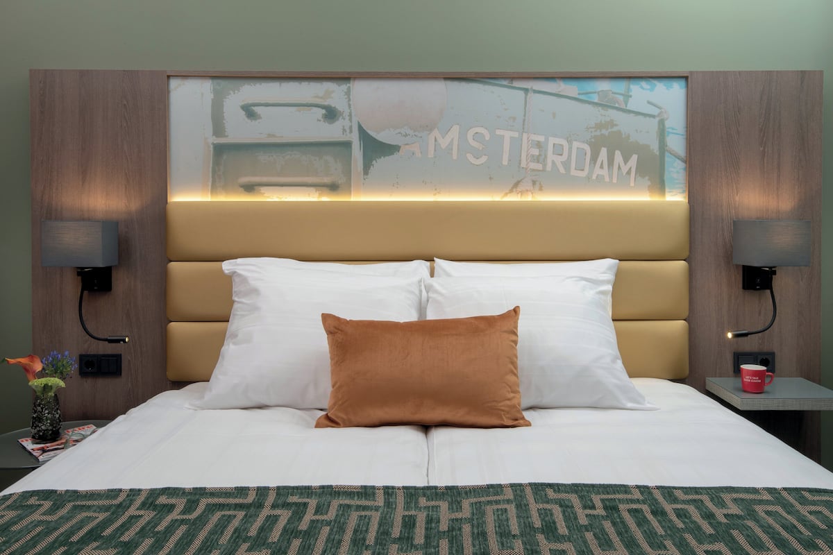 A hotel room with a large bed, two pillows, a nightstand with a lamp, a magazine, a vase with flowers, and a wall with a sign that says "AMSTERDAM".