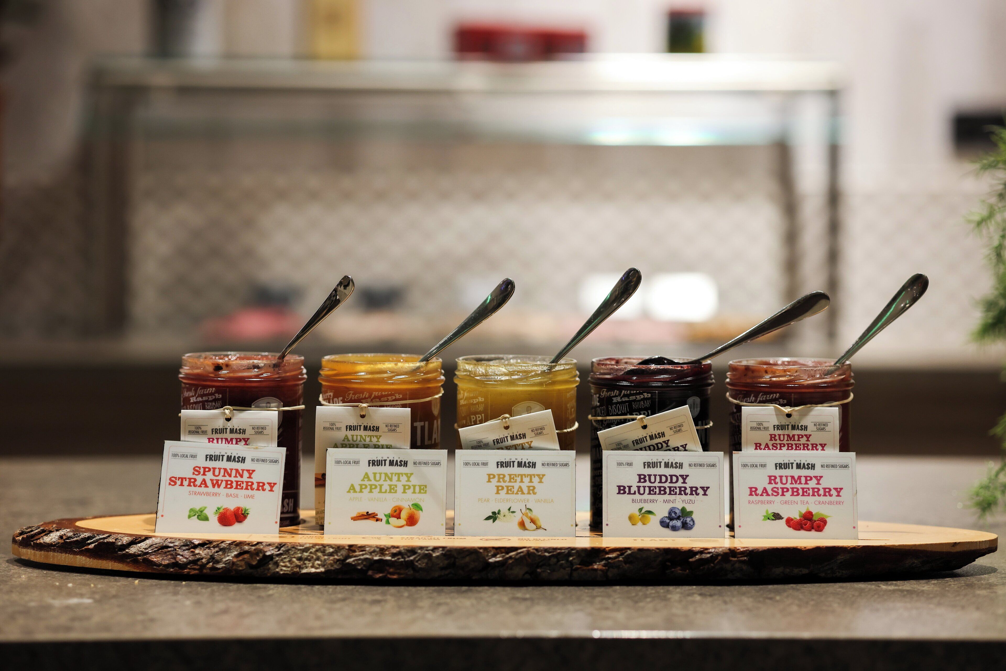 Variedad de conservas de frutas sobre una tabla de madera, con etiquetas que indican sabores como fresa, tarta de manzana, pera, arándano y frambuesa.