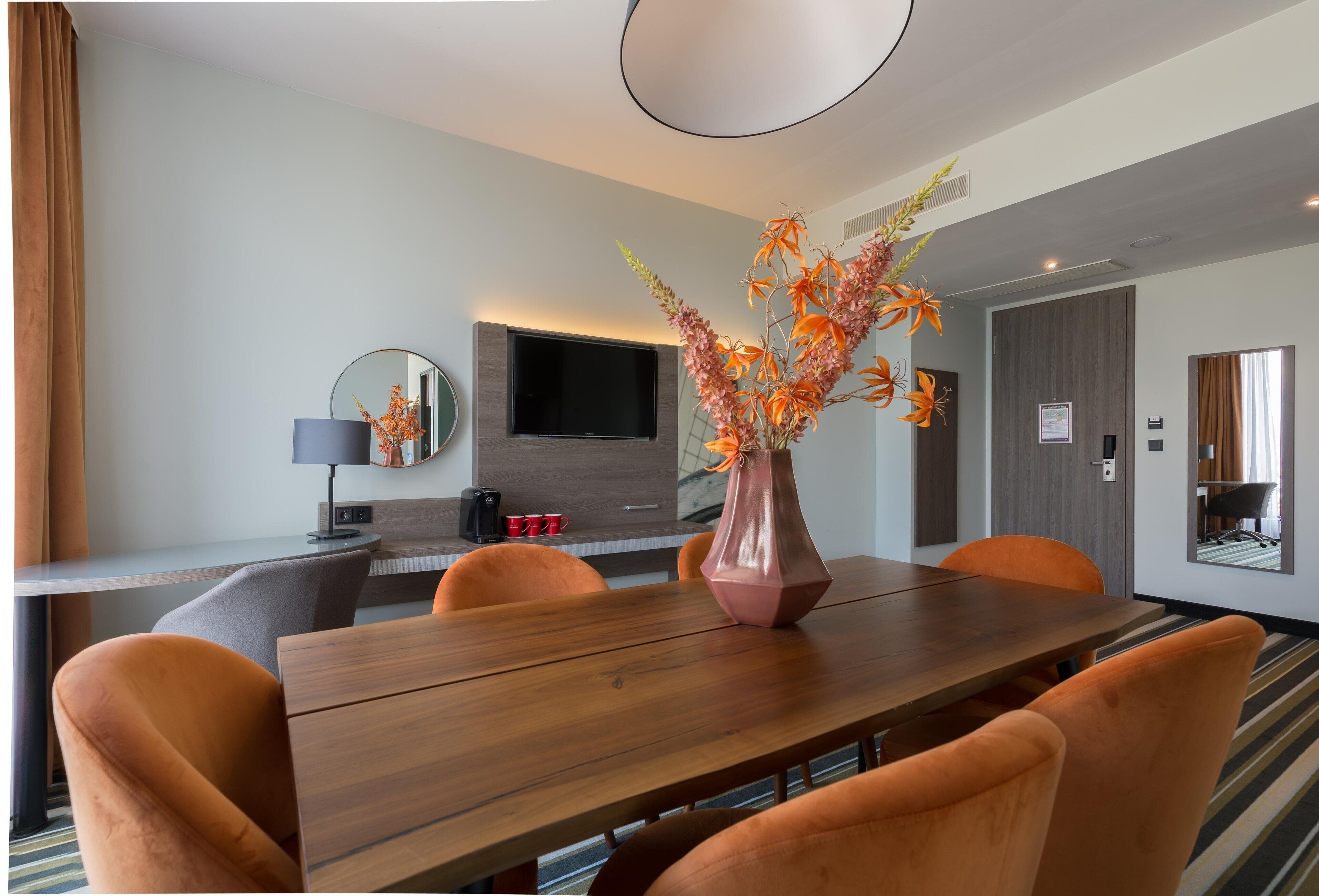 Una camera d'albergo moderna con un tavolo da pranzo in legno, sedie arancioni, uno specchio grande, una TV a schermo piatto e un vaso con fiori arancioni.
