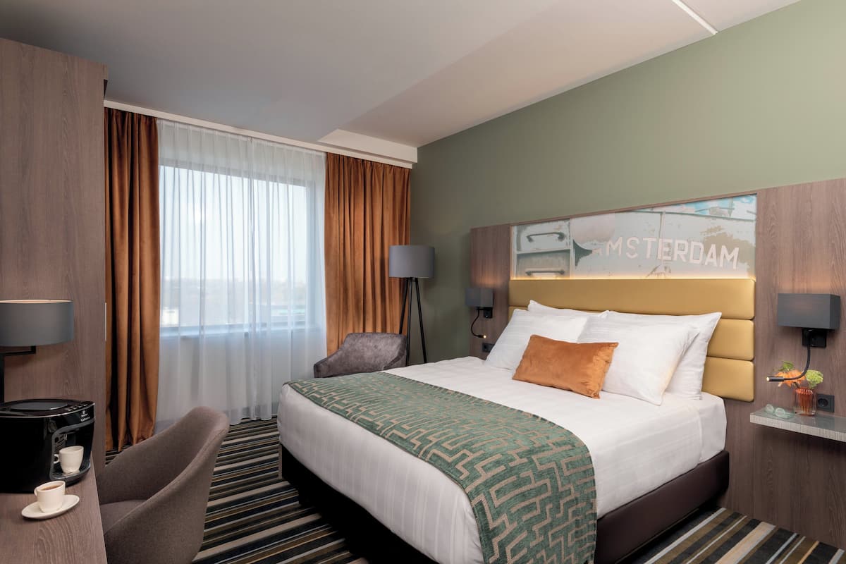 A hotel room with a large bed, a desk with a coffee machine, a chair, and a view of the city.