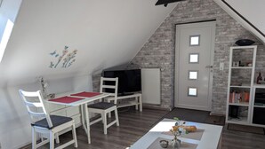 Quarto estilo loft com teto inclinado, mesa de jantar branca com cadeiras, televisão, estante de livros e radiador.