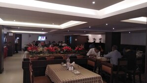 A restaurant with tables covered in patterned tablecloths, potted plants, and a television screen in the background. - Alhind Calicut Tower (Kozhikode)