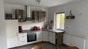 A modern kitchen with white cabinets, a built-in oven, a microwave, and a toaster. There is a window providing natural light and a bottle of wine on the dining table. - Ferienhaus Netzelkow am Achterwasser, Usedom, Baltic Sea, with WiFi, family-friendly (Lütow OT Netzelkow)
