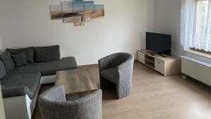 A modern living room with a grey sofa, two armchairs, a coffee table, and a flat-screen TV on a wooden stand. A multi-panel artwork is on the wall, and there's a window with sheer curtains. - Ferienhaus Netzelkow am Achterwasser, Usedom, Baltic Sea, with WiFi, family-friendly (Lütow OT Netzelkow)