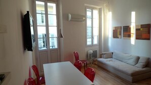 Una sala de estar con un sofá blanco, una mesa de comedor con sillas rojas y un televisor montado en la pared.
