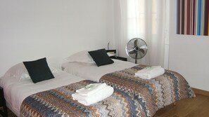 Habitación de hotel con dos camas, cada una con colcha y almohadas estampadas, un ventilador de pie y una ventana con cortinas.