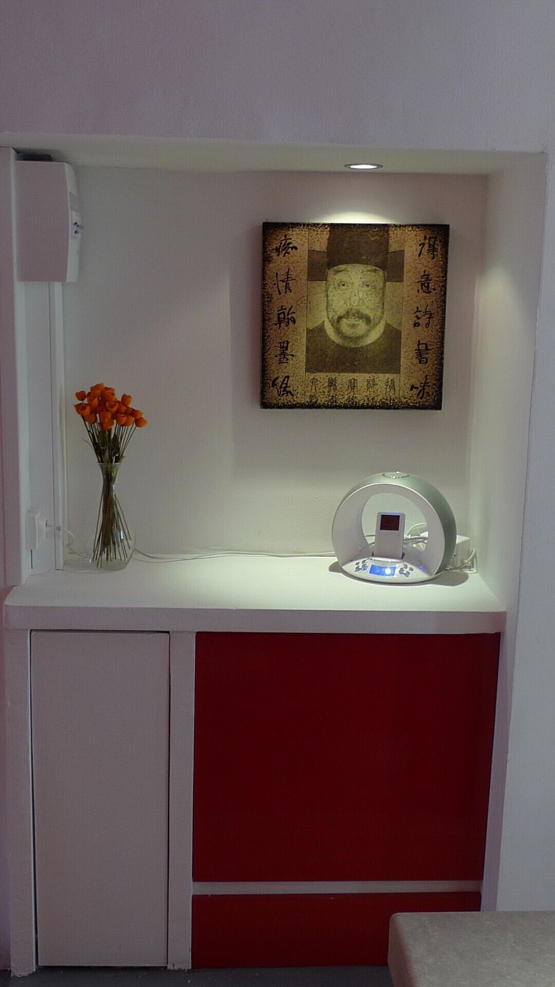 A small kitchenette with a white countertop, a vase with orange flowers, a framed picture, and a white appliance.
