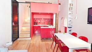 Una cocina moderna con armarios rojos y encimera blanca, una zona de comedor con mesa blanca y sillas rojas, y una escalera que sube a un nivel superior.