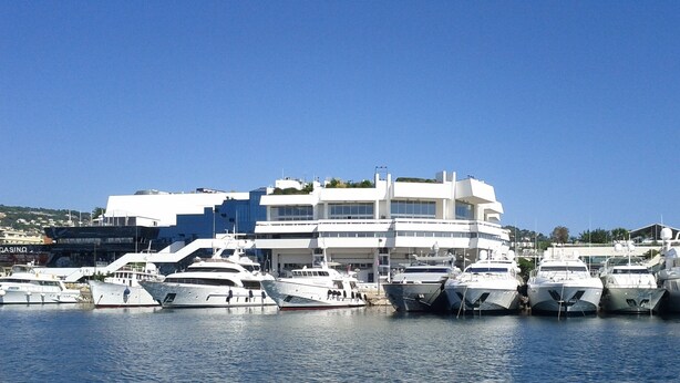 Un puerto deportivo con varios yates amarrados, un edificio blanco de varios pisos y un cielo azul despejado.
