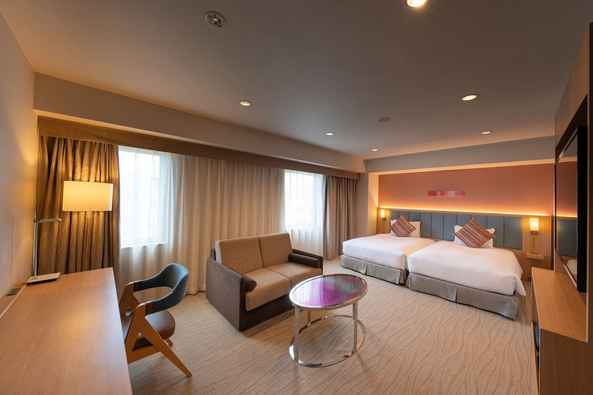 A hotel room with a sofa, two beds, a small table, and a lamp.