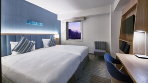 Habitación de hotel con una cama grande, un escritorio, una silla, un televisor y una ventana con vistas a la ciudad.