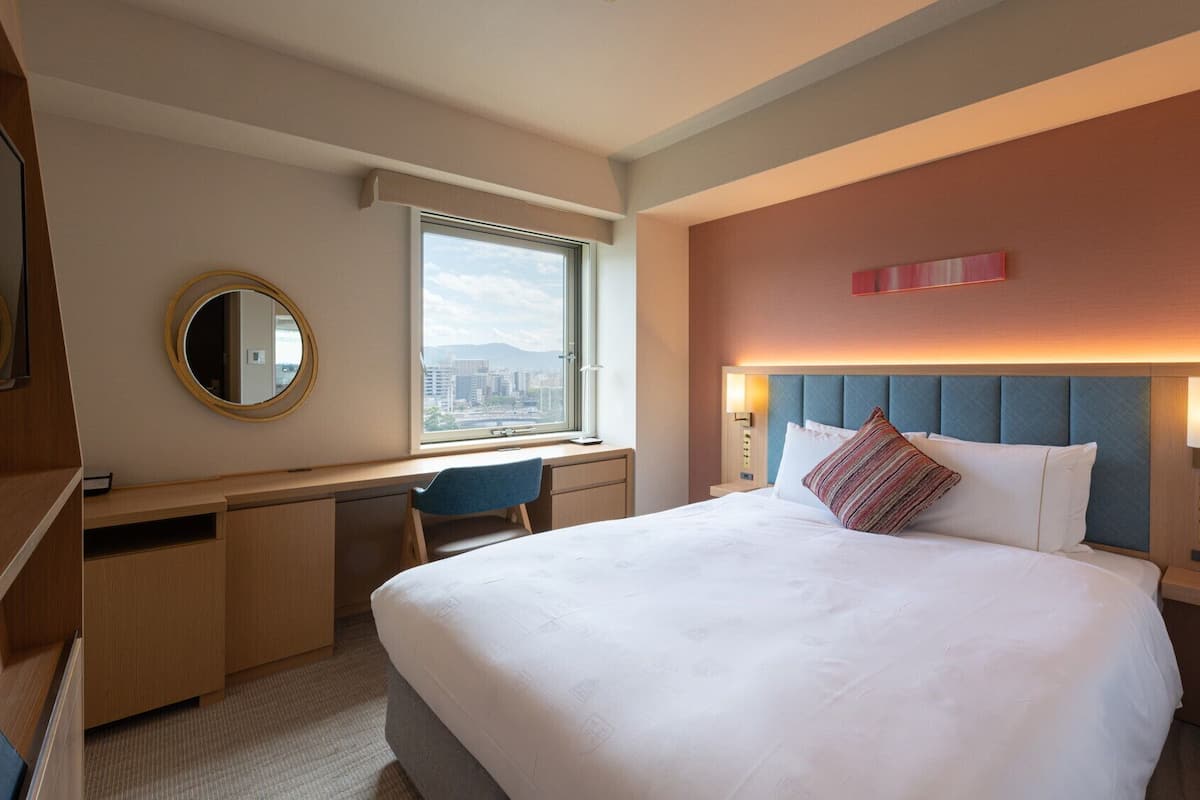 A hotel room with a bed, desk, chair, and a large window with a city view.