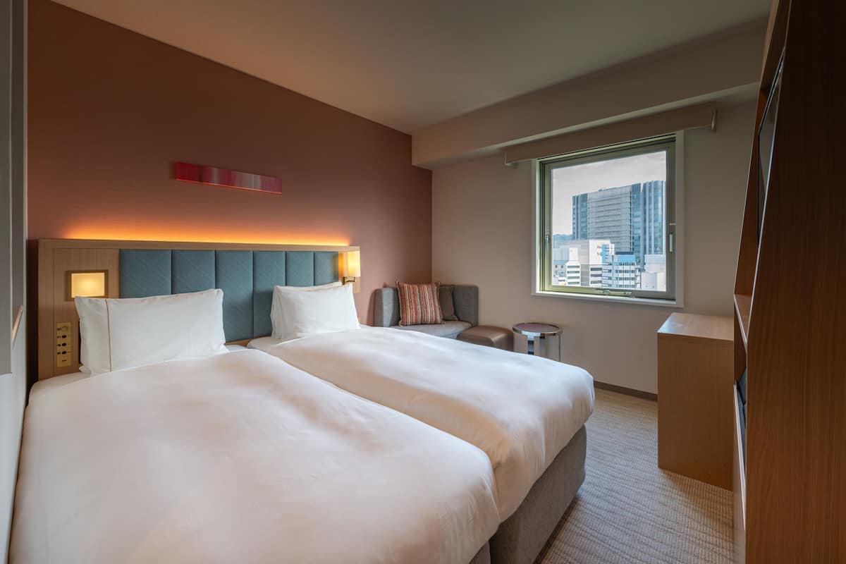 A hotel room with a large bed, a seating area, a small table, and a window with a city view.