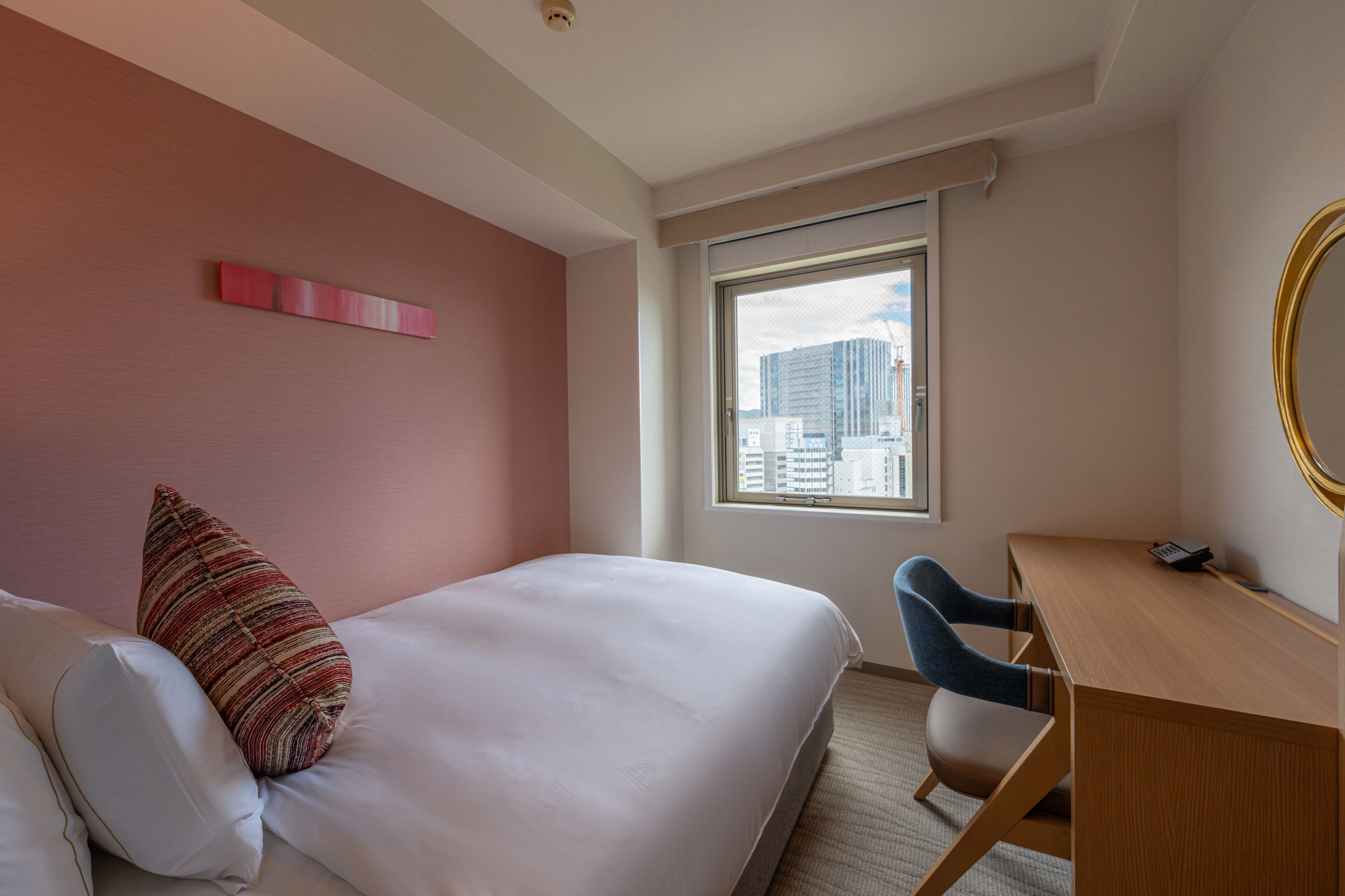A hotel room with a bed, a desk, a chair, a window with a view of buildings, and a wall-mounted mirror.