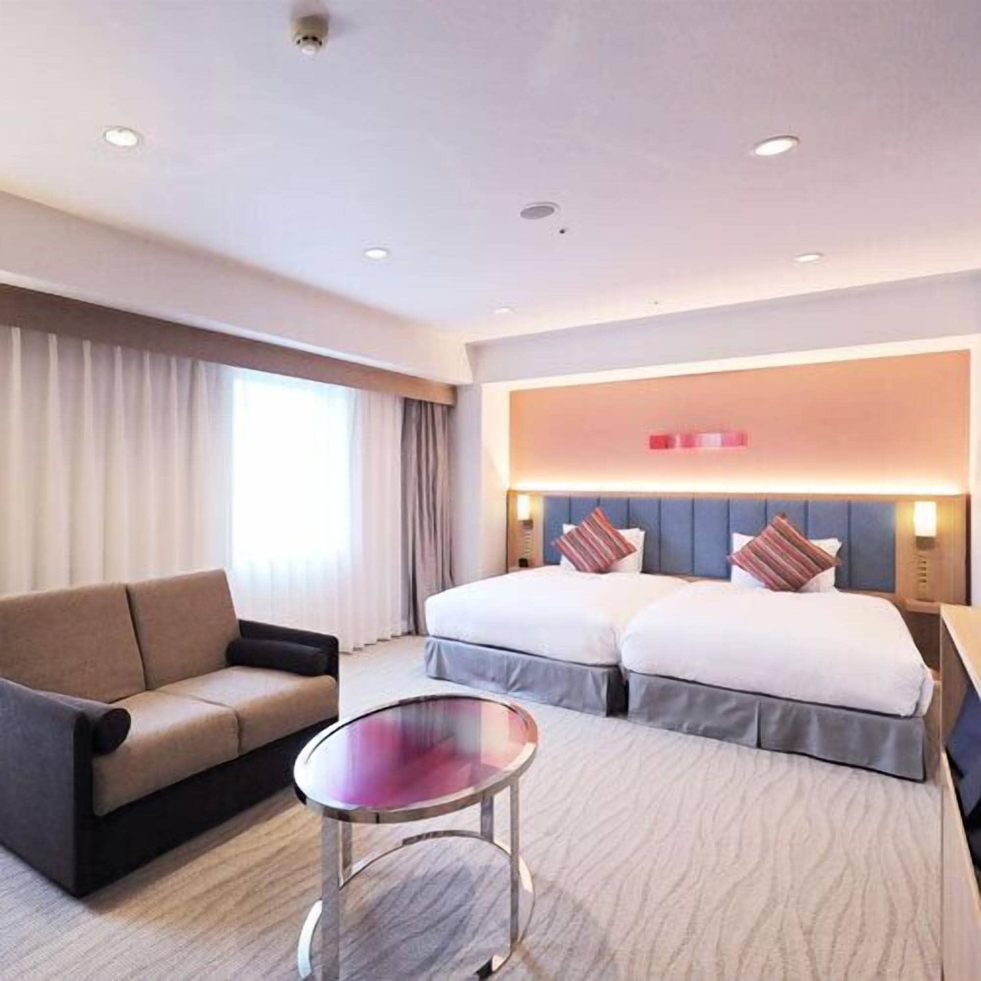 A hotel room with a sofa, two beds, a round table, and a large window with curtains.