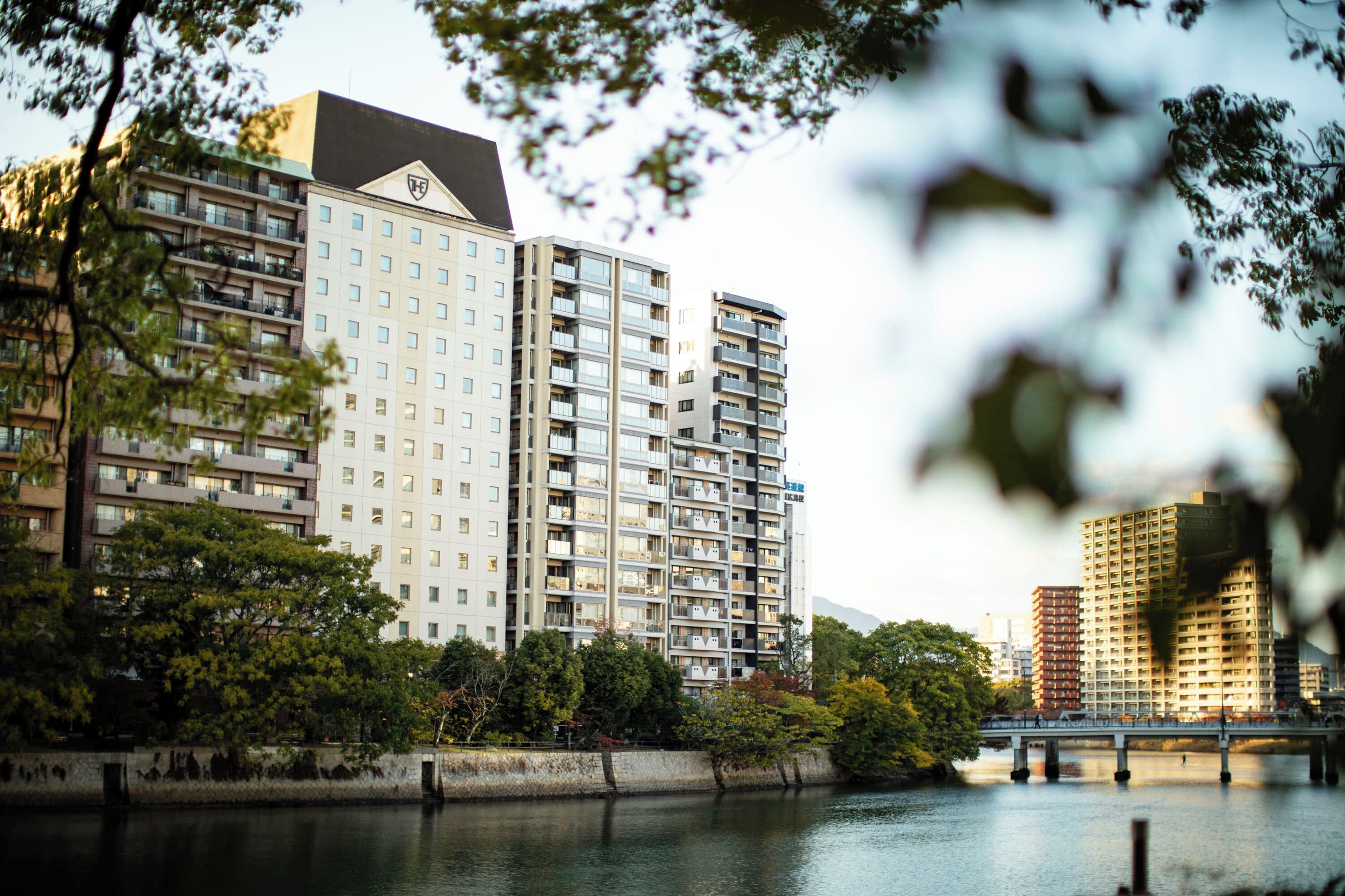 A multi-story hotel with balconies, a river in the foreground, and a bridge in the background.