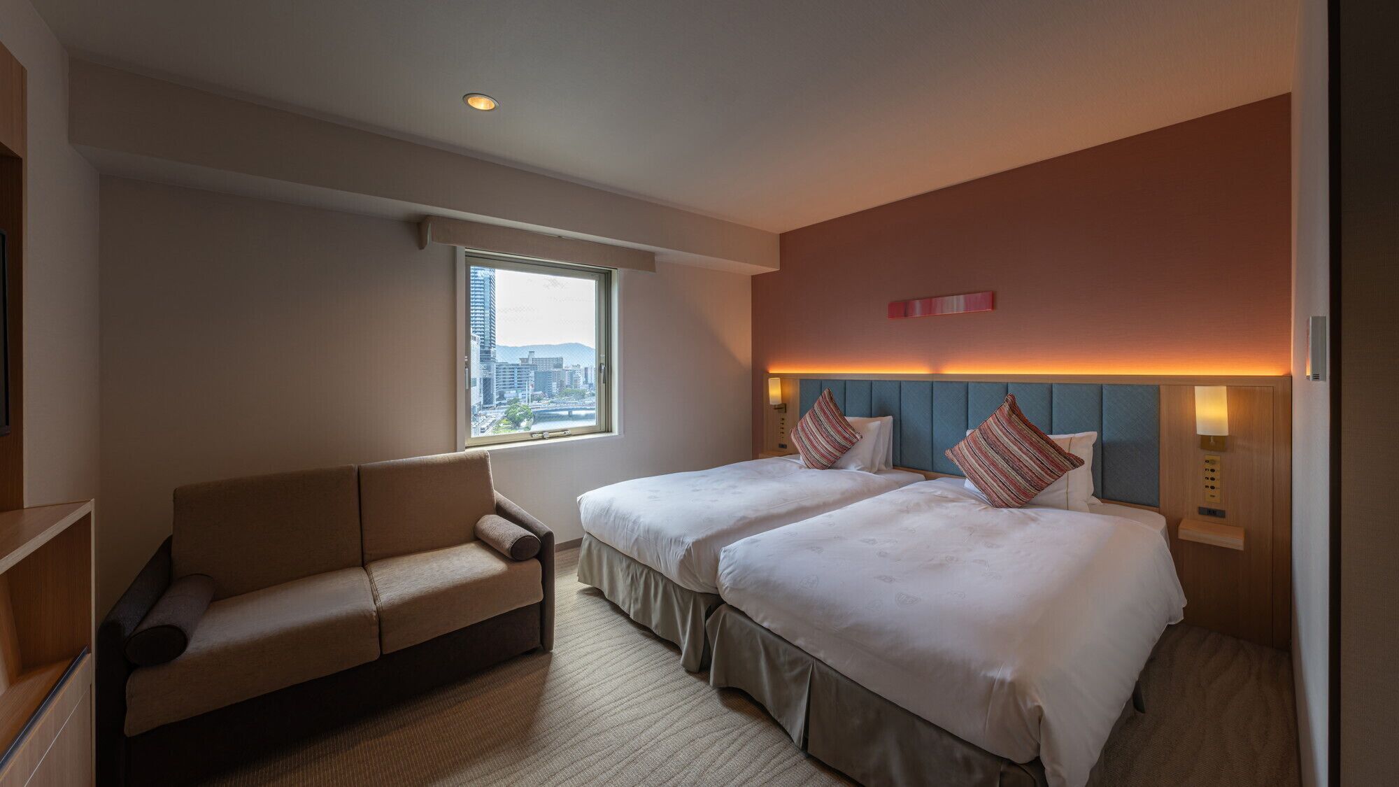 Habitación de hotel con dos camas, un sofá, una ventana con vista a la ciudad y una lámpara fijada a la pared.