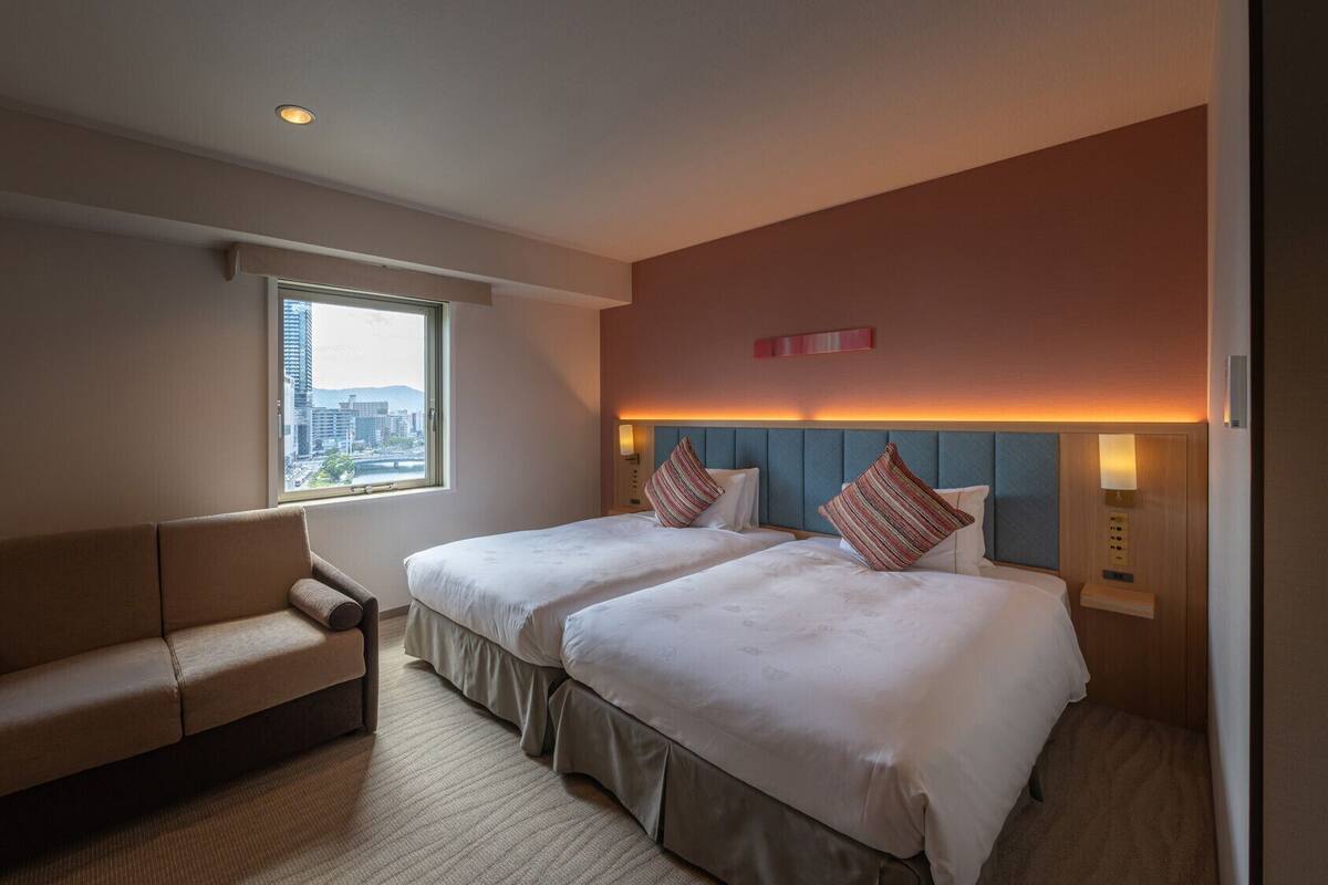 A hotel room with two beds, a sofa, a window with a city view, and a wall-mounted light fixture.