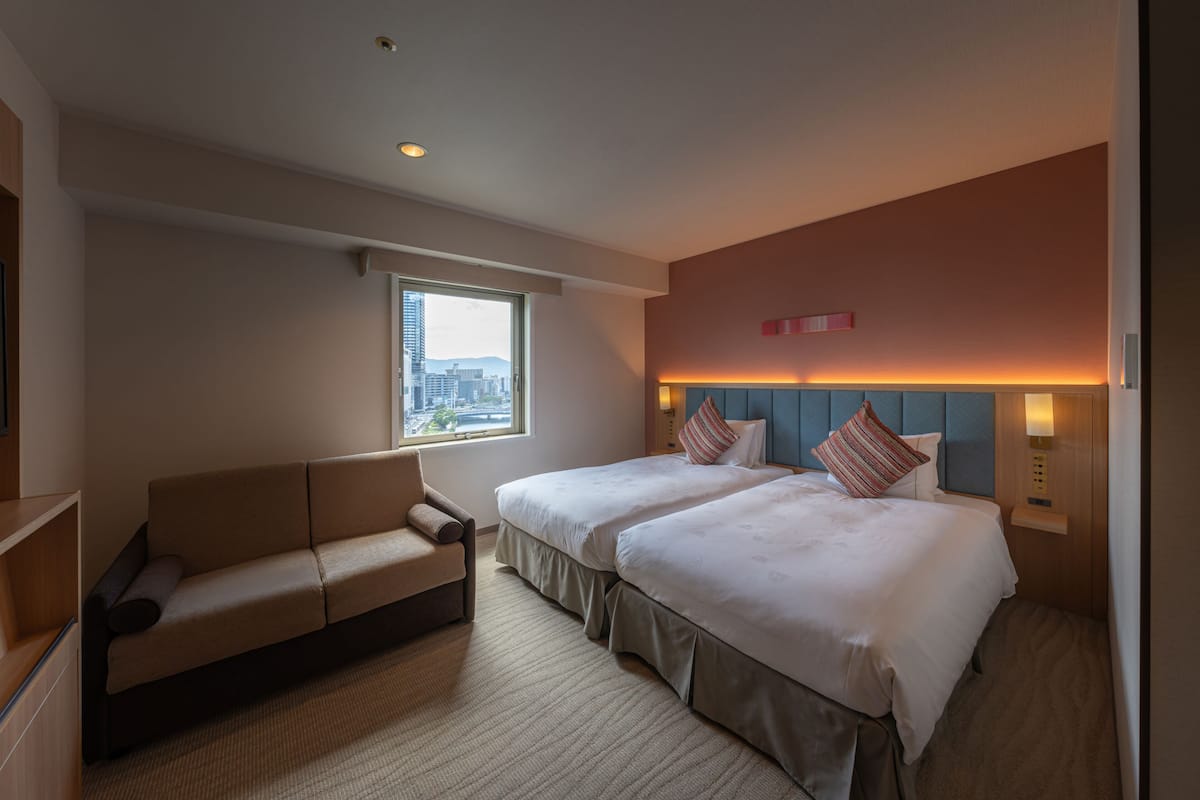 A hotel room with two beds, a sofa, a window with a city view, and a wall-mounted light fixture.