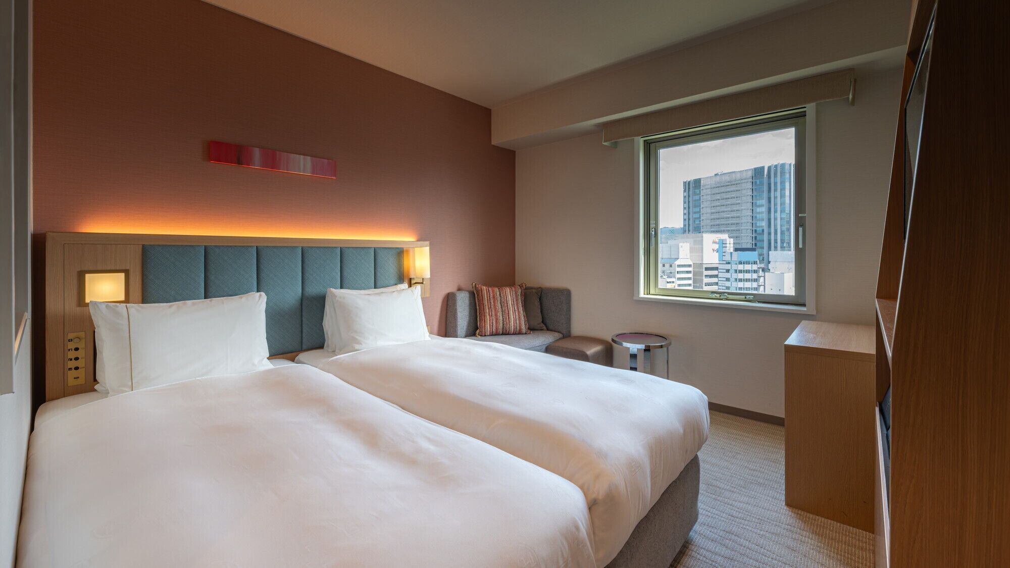 Habitación de hotel con una cama grande, cabecera con iluminación integrada, un pequeño rincón de descanso y una ventana con vista a la ciudad.