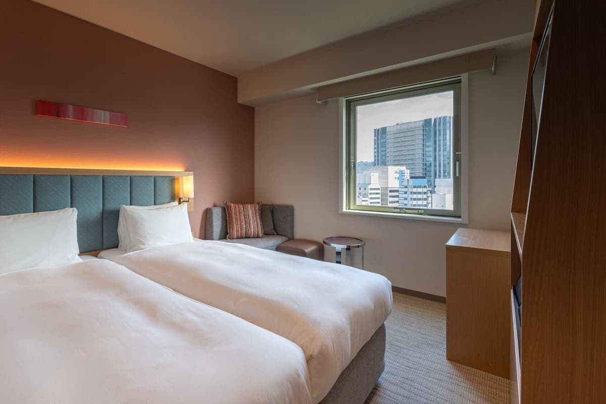 A hotel room with a large bed, a headboard with built-in lighting, a small seating area, and a window with a city view.