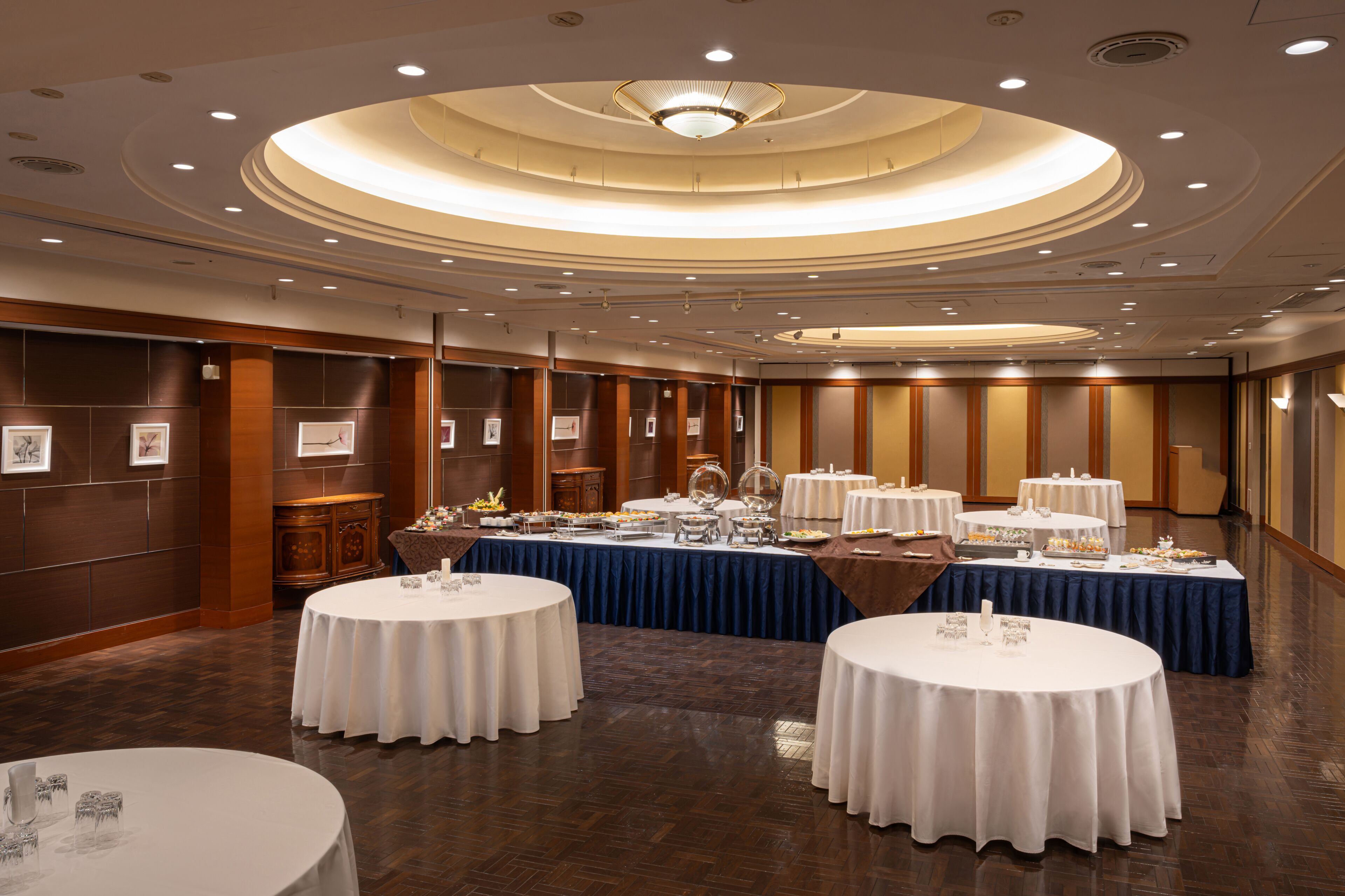 A banquet hall with round tables covered in white tablecloths, a central buffet area, and a circular ceiling design.