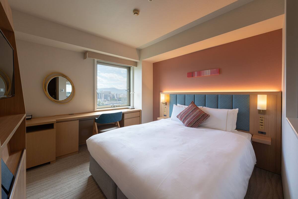 A hotel room with a large bed, a desk with a chair, a round mirror, a window with a city view, and a wall-mounted light fixture.