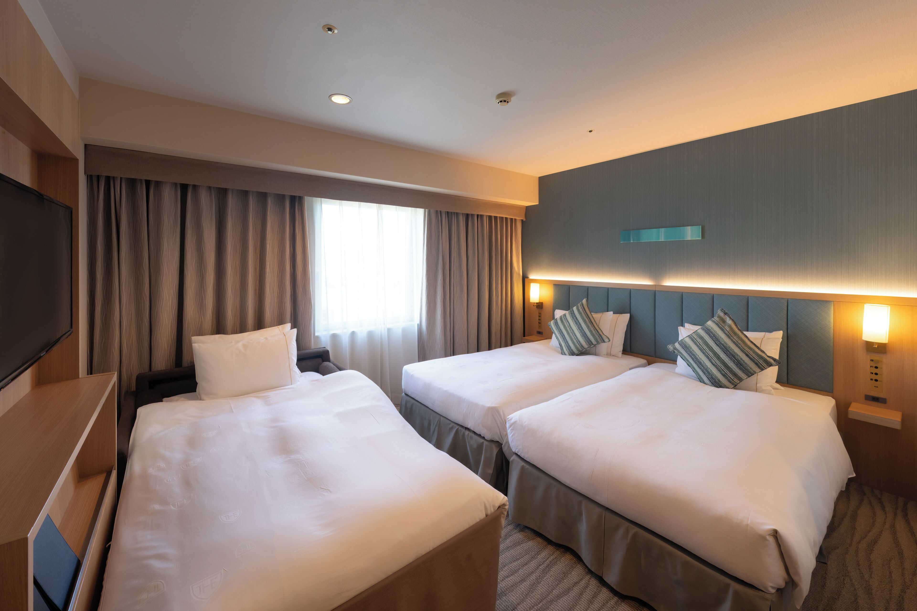 A hotel room with two beds, a TV, a window with curtains, and a wall-mounted light fixture.