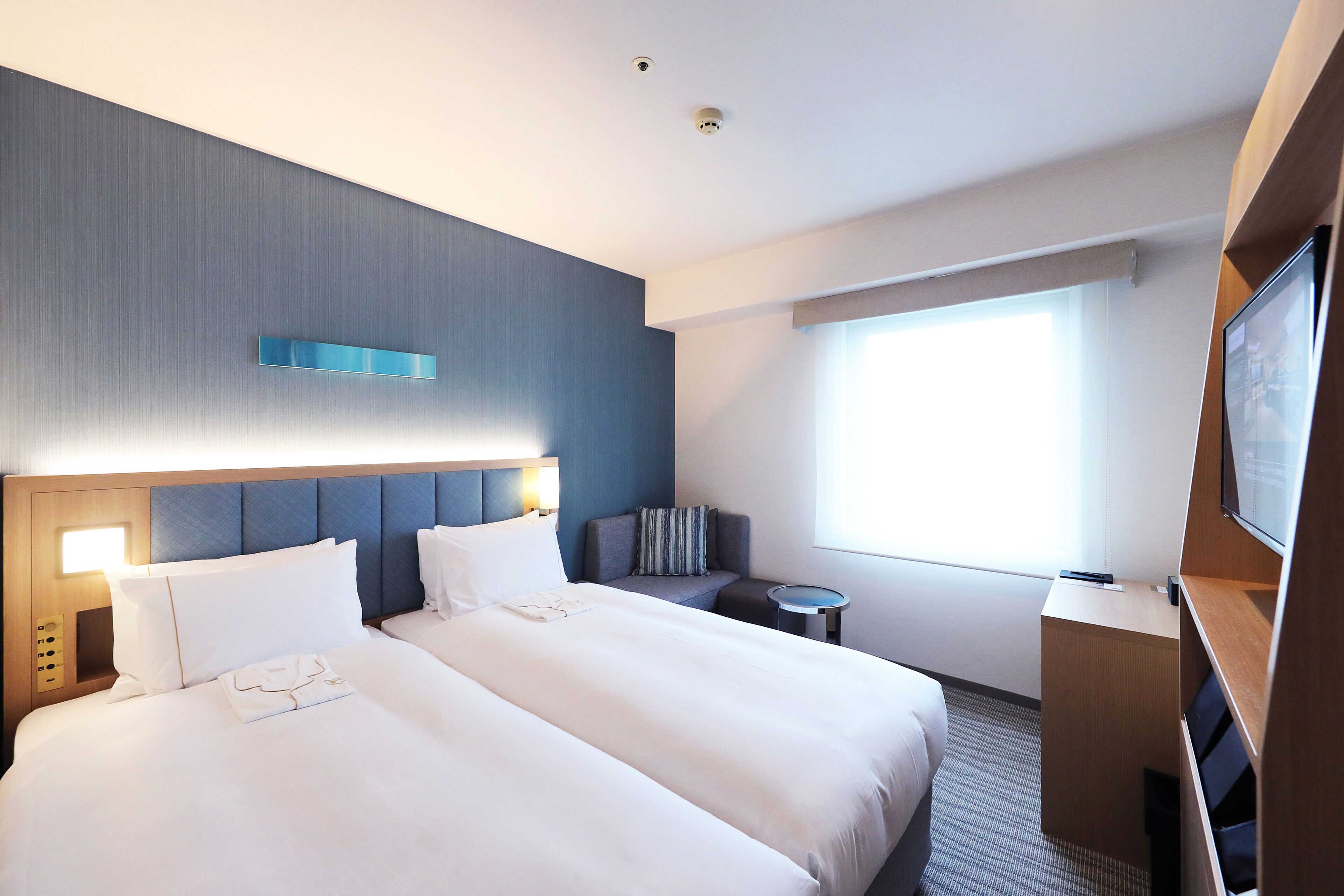 A hotel room with two beds, a desk, a chair, a TV, and a window with curtains.
